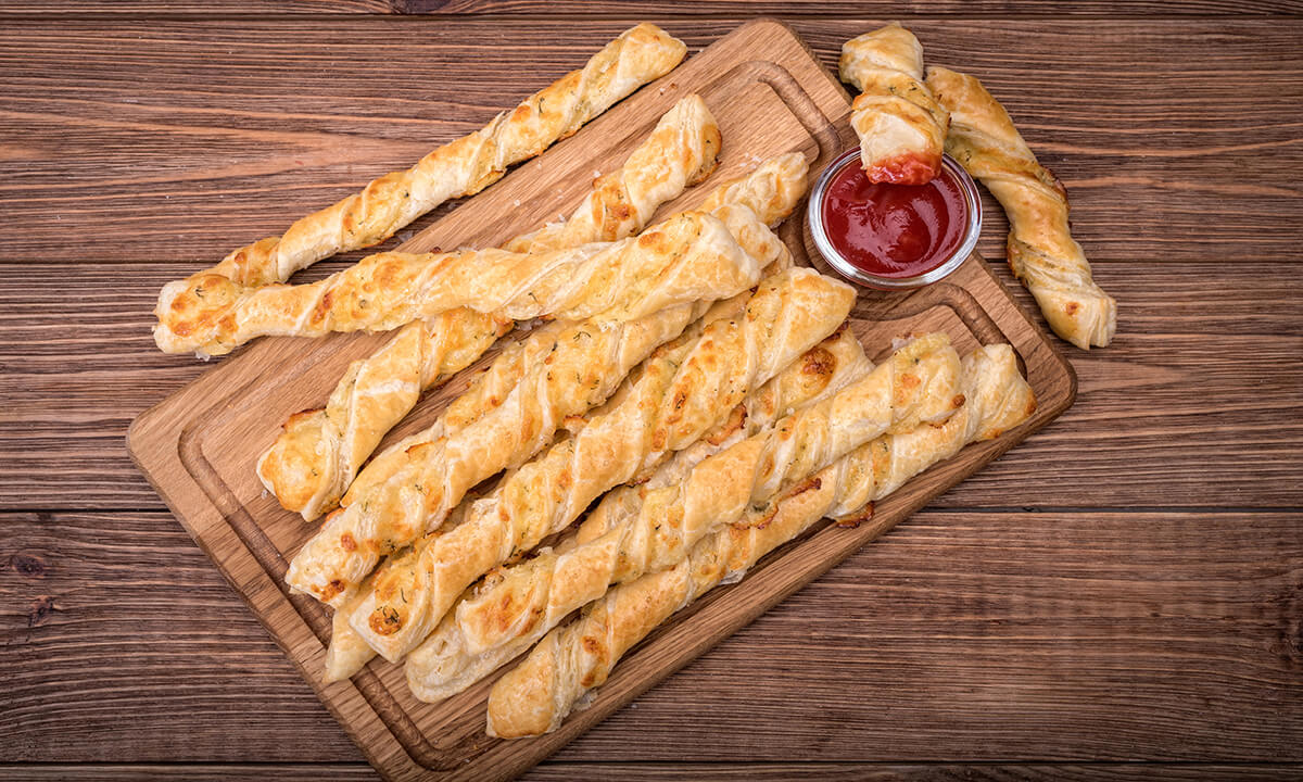 Palitos de queso sobre tabla de madera con salsa de tomate en un recipiente.