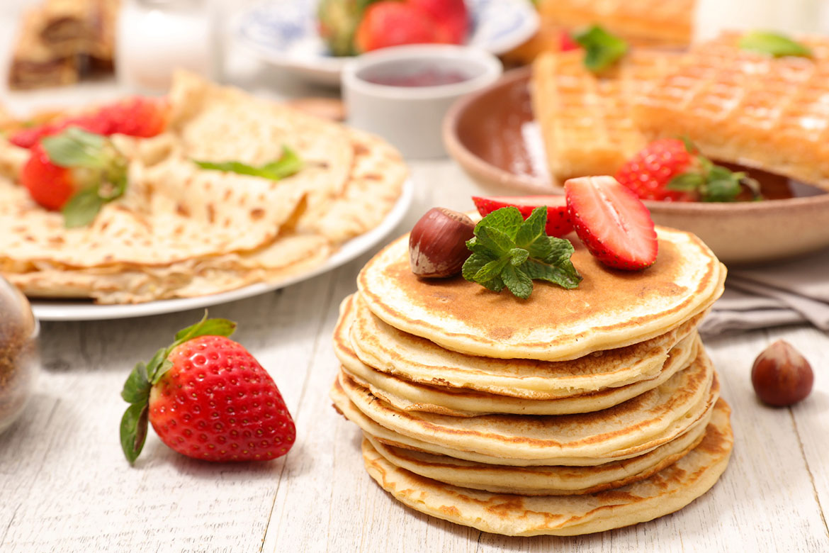 Pancakes apilados con fresas y hojas de menta en primer plano, acompañados de waffles y crepes sobre una mesa.