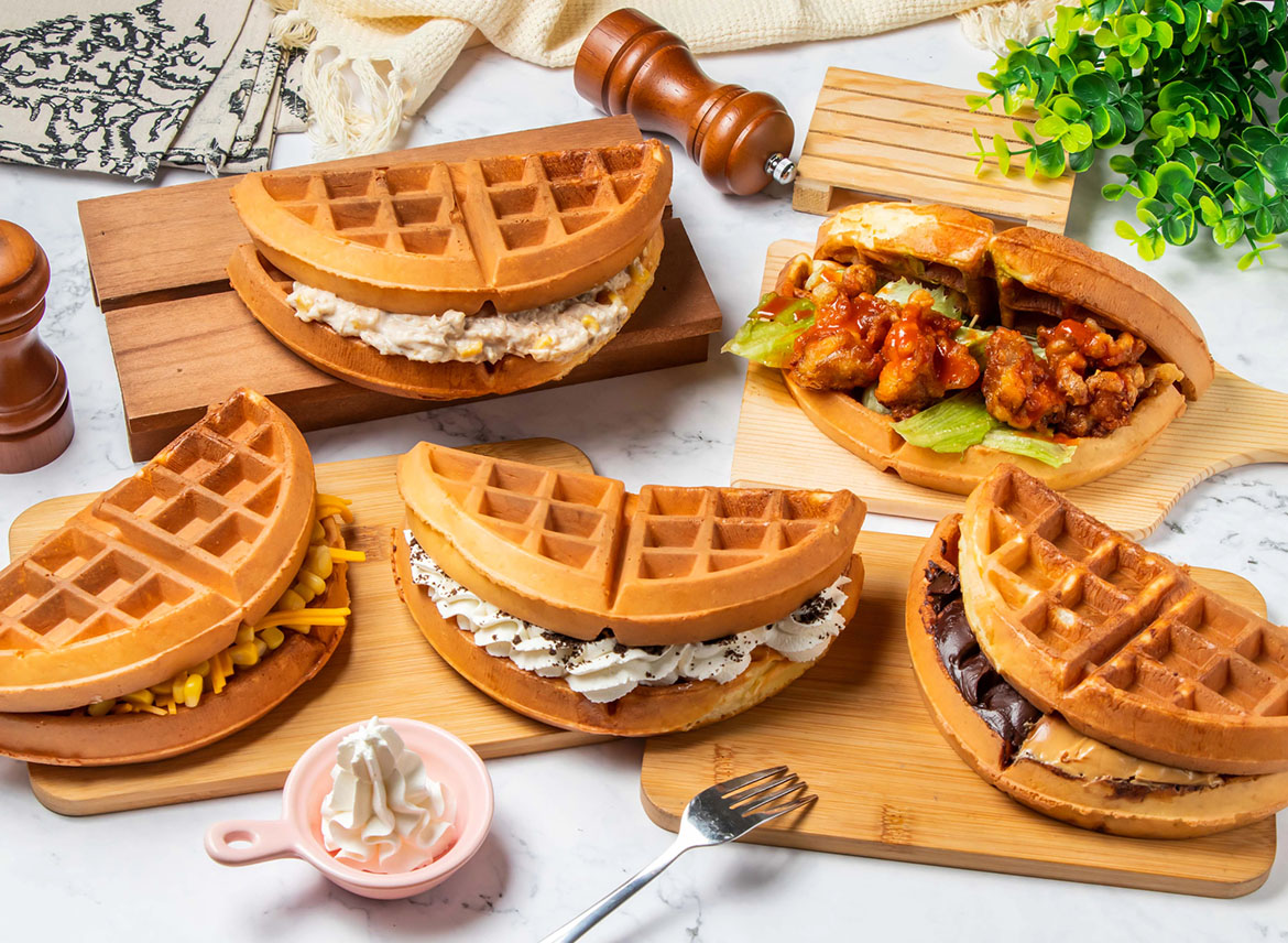 Variedad de waffles dulces y salados rellenos con chocolate, crema pastelera, queso, maíz y pollo sobre tablas de madera.