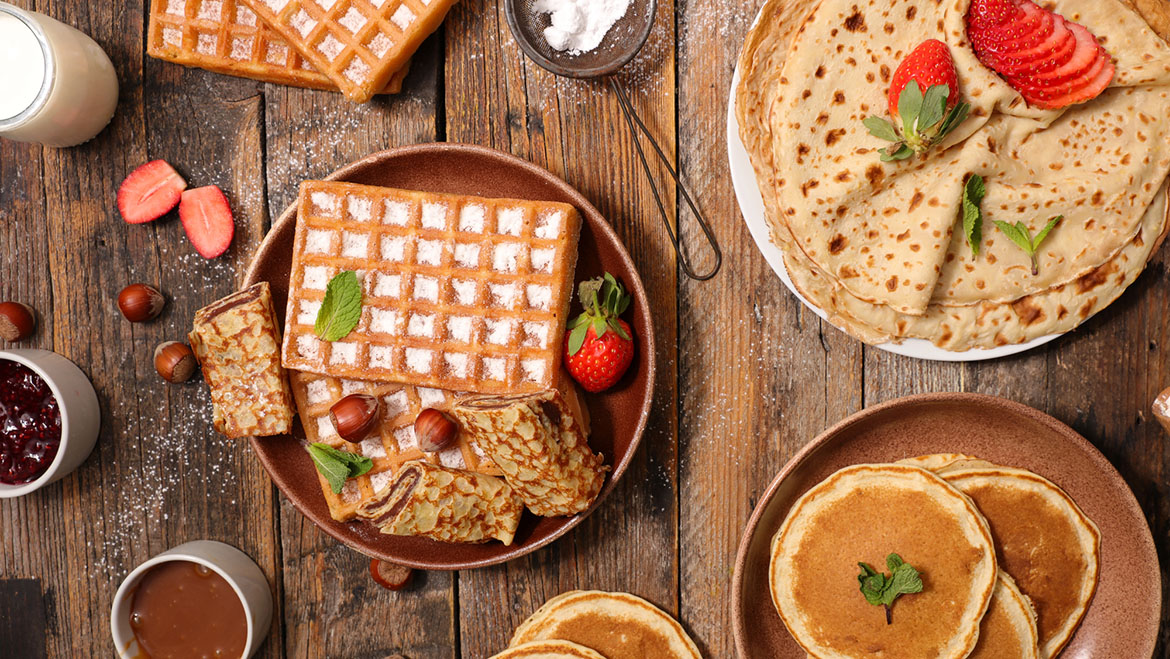 affles, pancakes y crepes servidos con frutas, fresas y acompañamientos sobre una mesa de madera.
